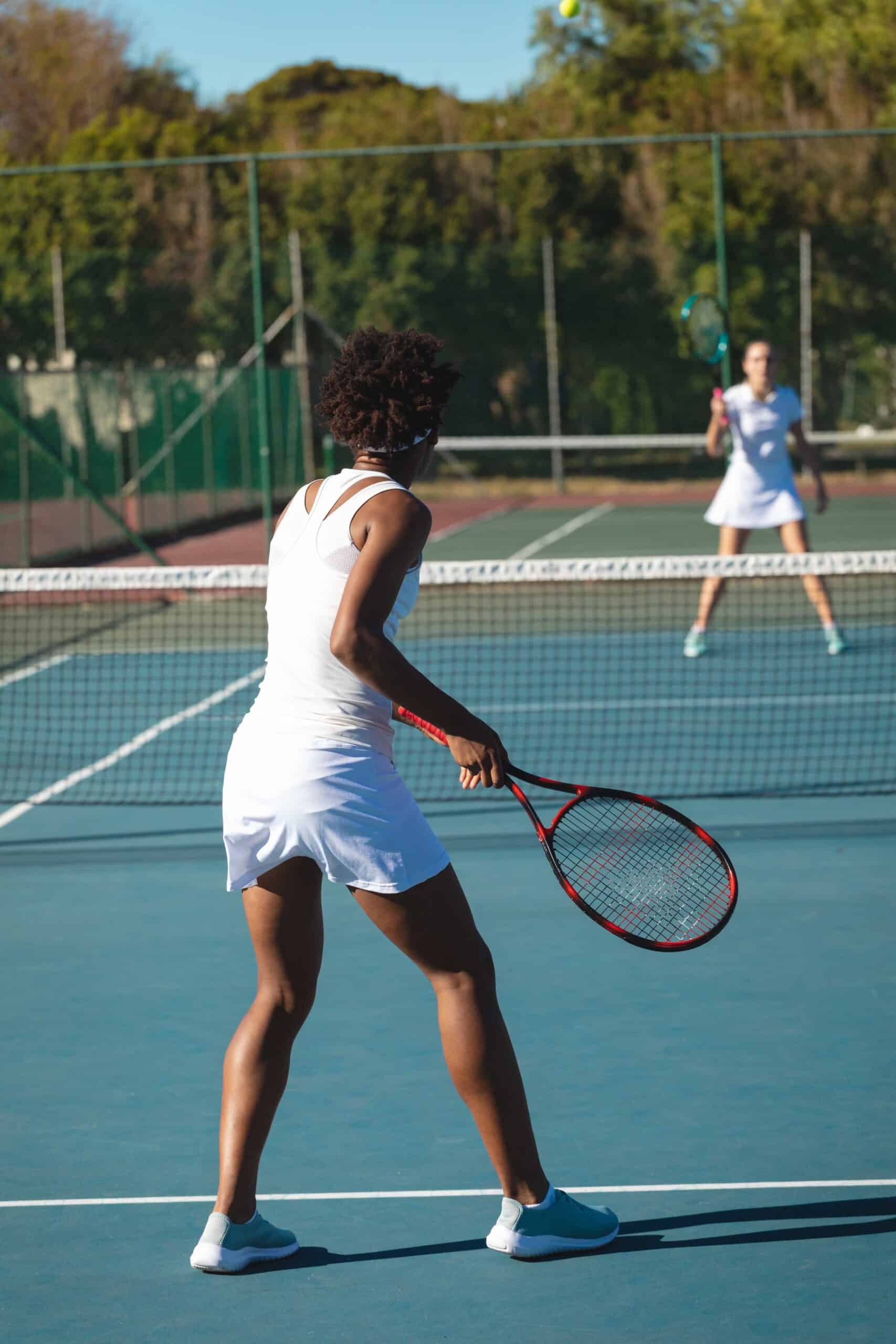 Zwei Tennisspielerinnen in weißen Outfits spielen ein Match auf einem Tennisplatz bei sonnigem Wetter.