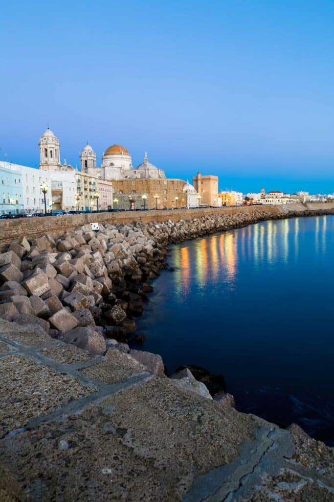 Uferpromenade mit einer Kathedrale in Cádiz, Spanien, bei Sonnenuntergang, mit reflektierenden Lichtern im Wasser.
