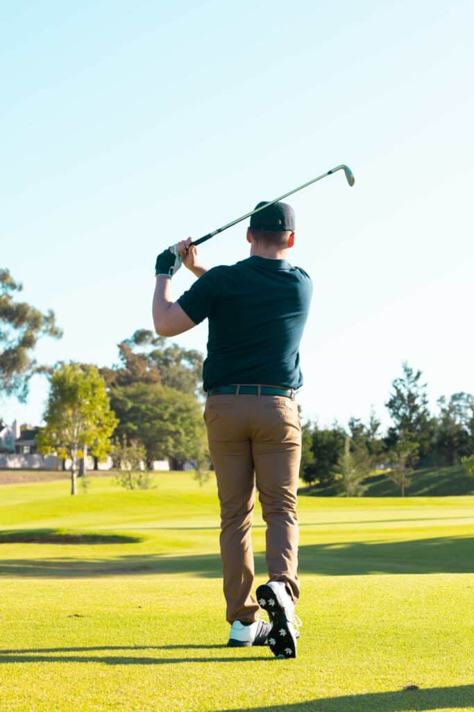Ein Golfer in einem dunklen Shirt und braunen Hosen steht auf einem grünen Golfplatz und führt einen Schwung mit einem Golfschläger aus, umgeben von Bäumen und klarem Himmel.