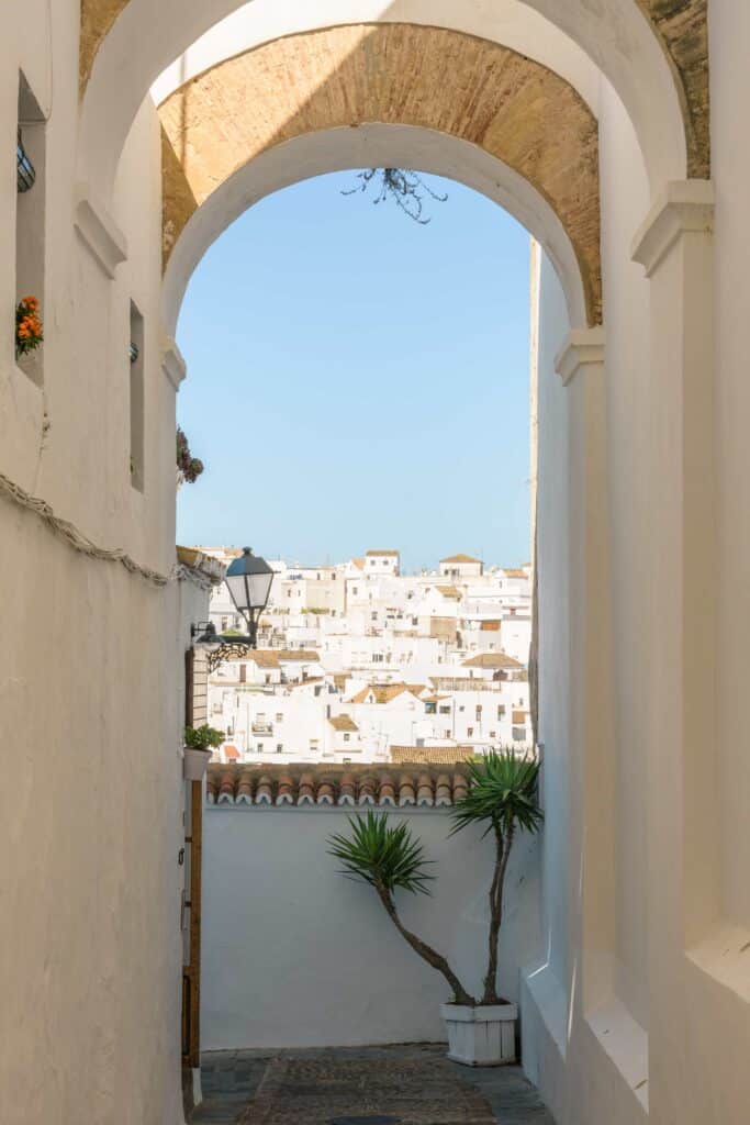 Ein weißer, überdachter Durchgang führt in ein Dorf mit weißen Häusern. Im Vordergrund steht eine Laterne und ein Topf mit einer grünen Pflanze, während hellblauer Himmel die Szene erhellt.