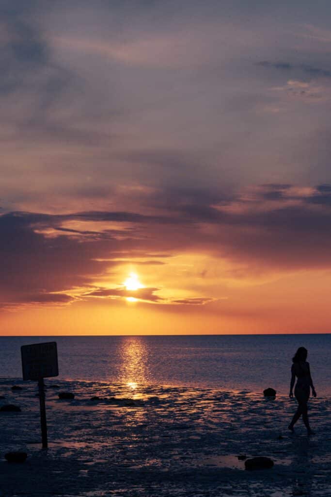 Silhouette einer Person, die bei Sonnenuntergang am Strand entlangläuft, mit einem orangefarbenen Himmel und reflektierendem Wasser im Hintergrund.
