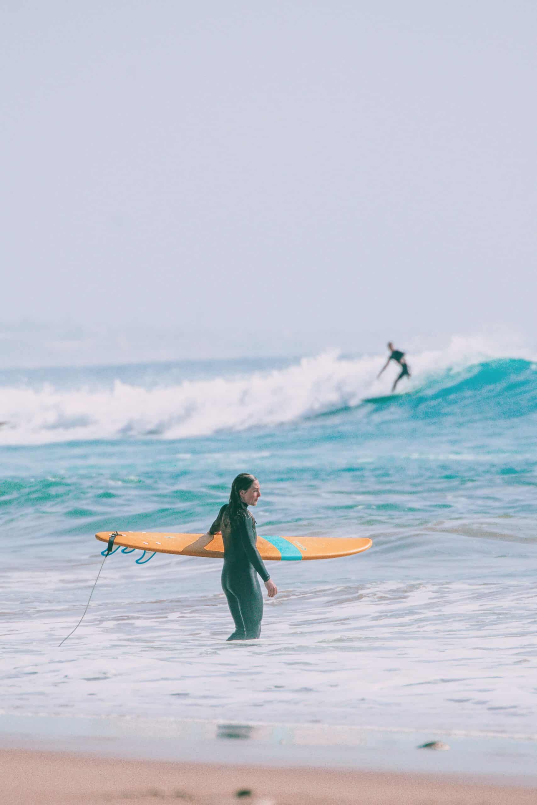 Eine Person in einem Neoprenanzug steht mit einem großen gelben Surfbrett im Meer, während eine andere Person auf einer Welle im Hintergrund surft.