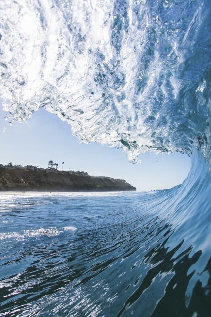 Eine große, durchscheinende Meereswelle rollt auf einen felsigen Küstenabschnitt zu, während die Sonne auf das klare blaue Wasser scheint.