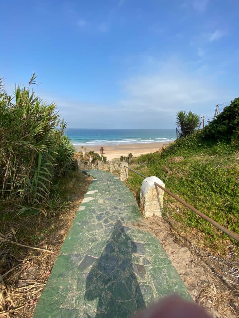 Ein steiniger Weg führt durch grüne Vegetation hinunter zu einem breiten Sandstrand und dem Meer, über dem ein blauer Himmel mit einigen Wolken schwebt.