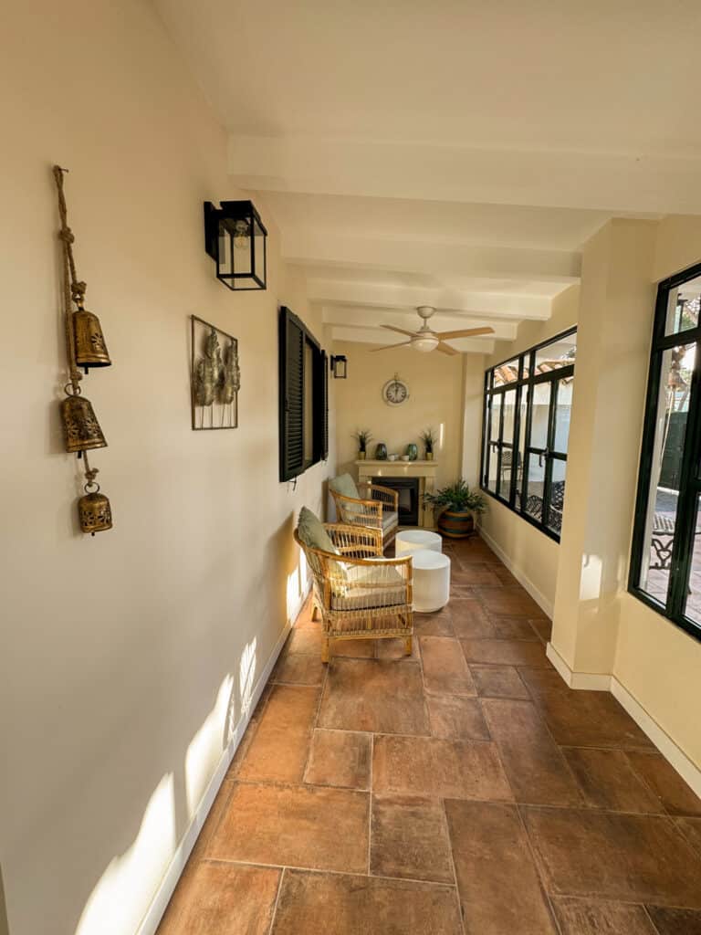 Ein heller Flur mit Terrakottafliesen, links mit einer Wanddekoration und zwei Laternenleuchten. Rechts stehen zwei geflochtene Stühle mit Kissen vor großen Fenstern. Im Hintergrund befindet sich eine Uhr über einem kleinen Tisch mit Pflanzen und Dekorartikeln.
