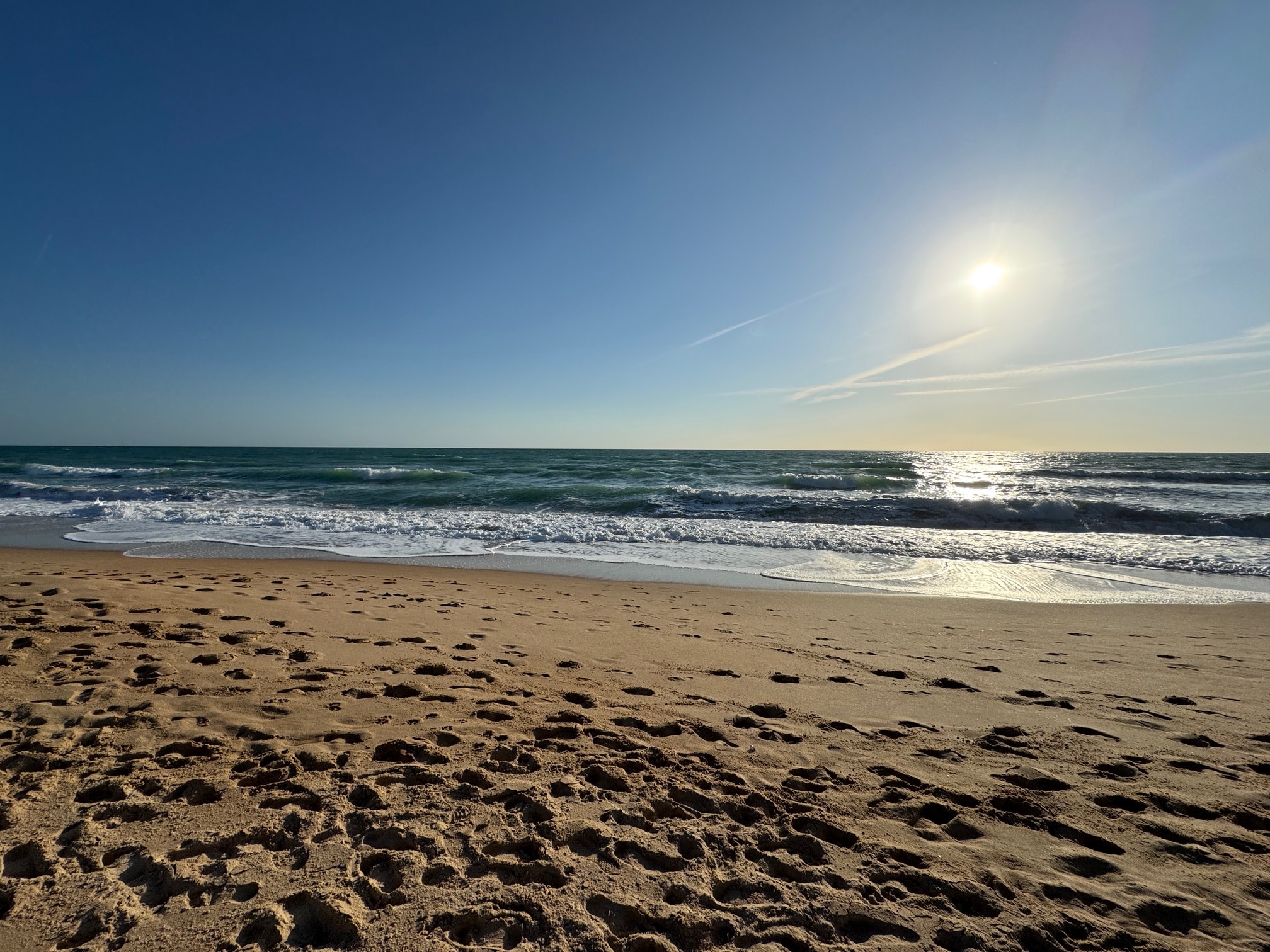 Sonniger Strand mit Fußabdrücken im Sand, Wellen, die an das Ufer schlagen, und klarem blauen Himmel.