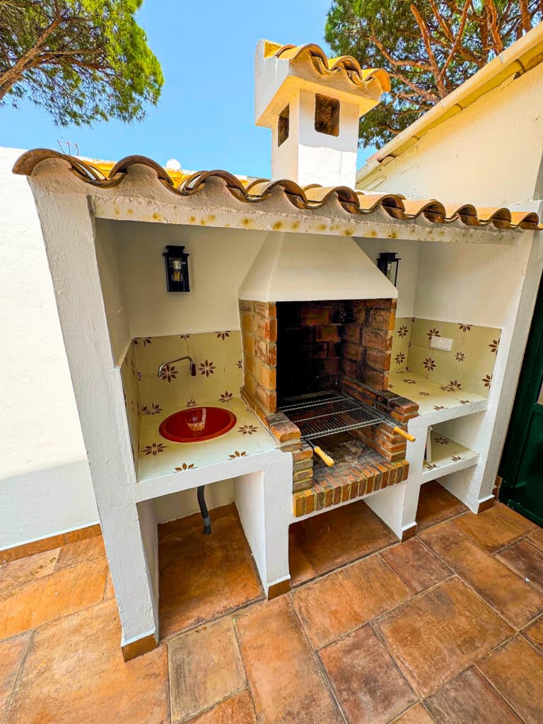 Ein gemauerter Außengrill mit Ziegeldach und integrierter Spüle, dekoriert mit geblümten Fliesen neben einem weißen Putzgebäude, umgeben von Terrakottafliesen und Bäumen im Hintergrund.