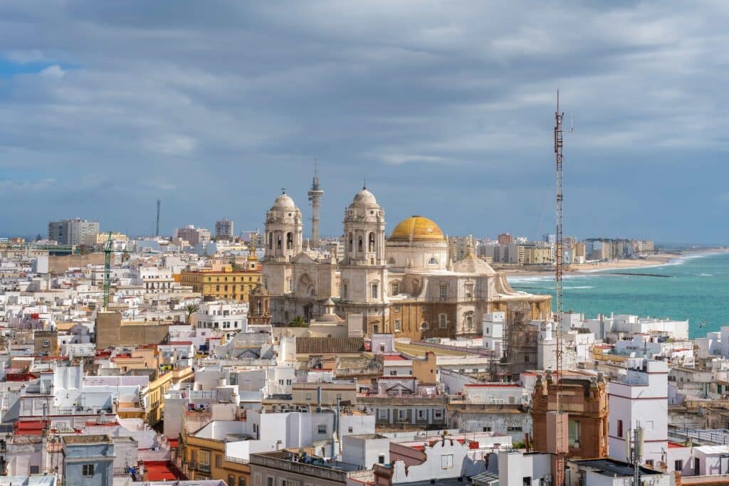 Panoramablick auf die Stadt Cádiz, Spanien, mit der Kathedrale von Cádiz im Vordergrund und dem Ozean im Hintergrund unter einem bewölkten Himmel.