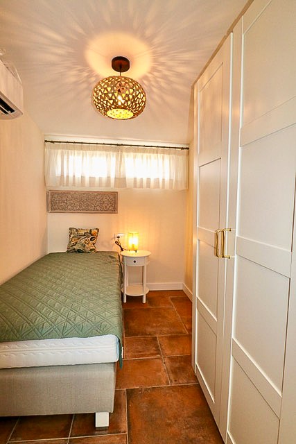 Kleines Schlafzimmer mit einem Einzelbett, das einen grünen Bettüberwurf hat, neben einem weißen Nachttisch mit Lampe. Ein weißer Schrank steht auf der rechten Seite, und eine dekorative Lampe hängt von der Decke. Der Raum hat Terrakottafliesen und eine kleine Fensteröffnung mit weißen Vorhängen.